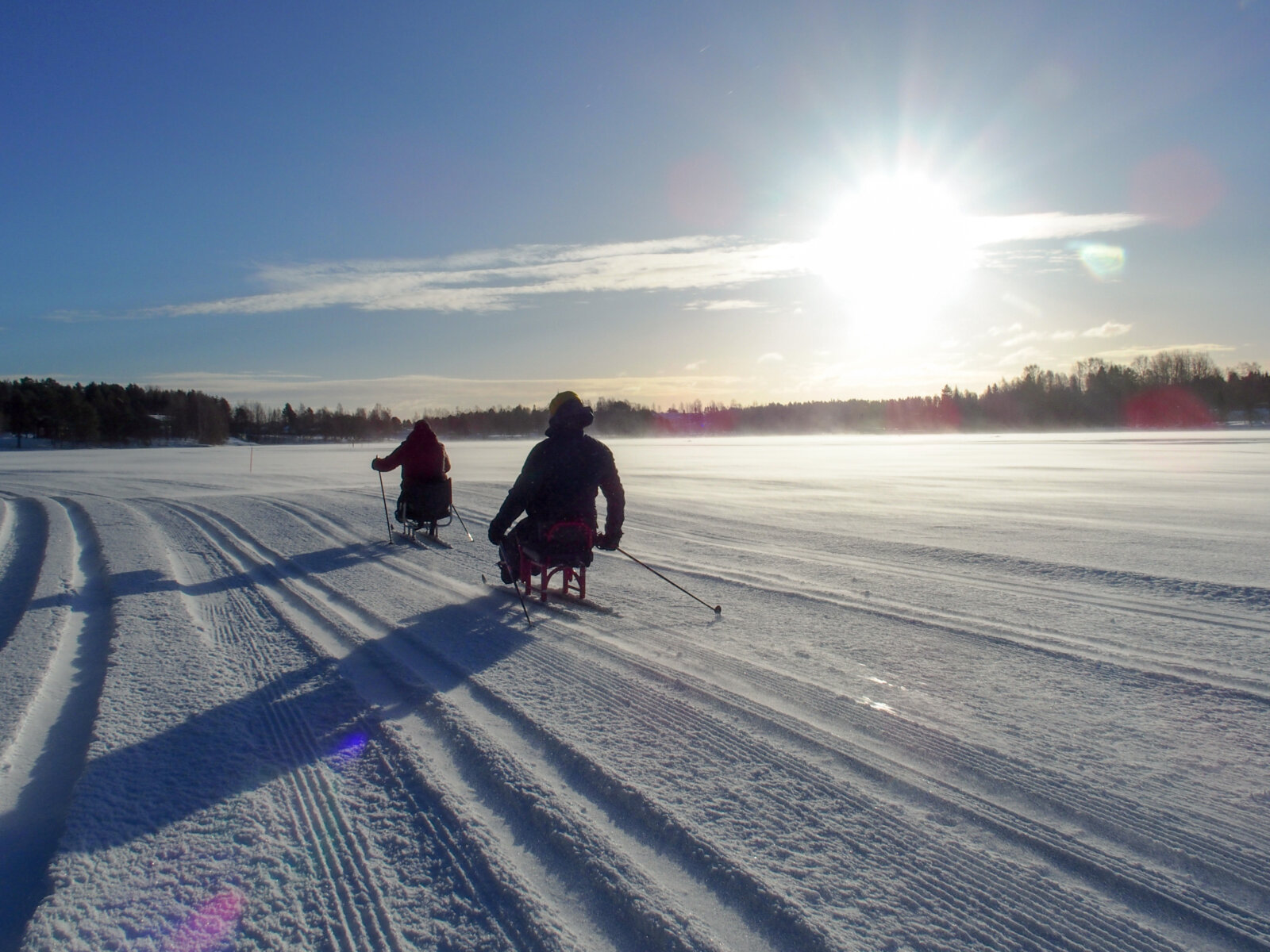 Kaksi kelkkahiihtäjää hiihtää pois päin kamerasta aurikoisella järviladulla.